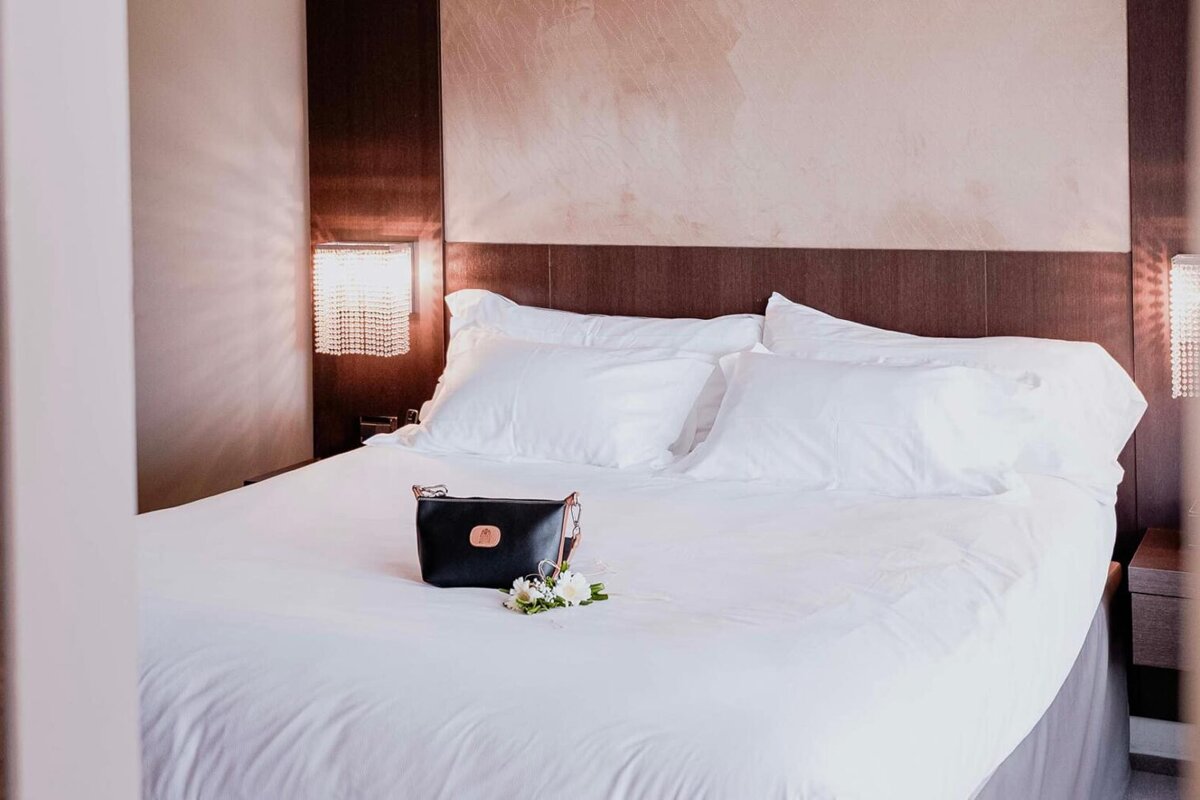 A bed with white sheets and pillows and a black bag on it