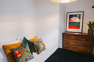 A cozy bedroom with a bed featuring teal, yellow, and floral pillows, a wooden dresser with abstract art, a plant, and white walls.