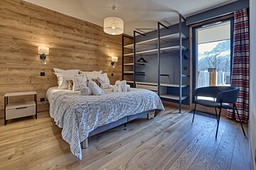 Stylish bedroom with a wooden accent wall, a comfortable bed, an open wardrobe, and a balcony showcasing a snowy mountain view.