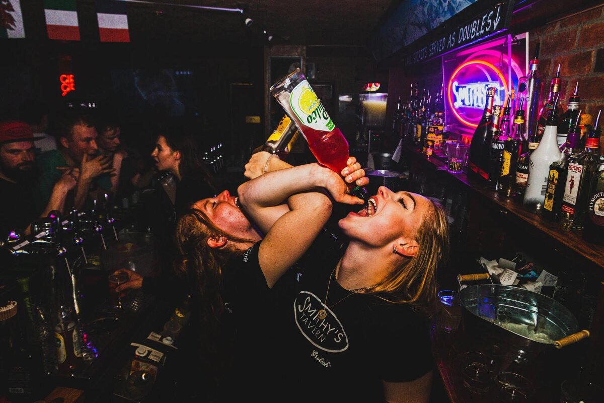 Two women in a dark, lively bar, one drinking directly from a red bottle while another's arm is wrapped around her. Neon lights and patrons visible.