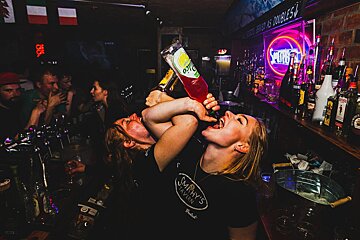 Two women in a dark, lively bar, one drinking directly from a red bottle while another's arm is wrapped around her. Neon lights and patrons visible.