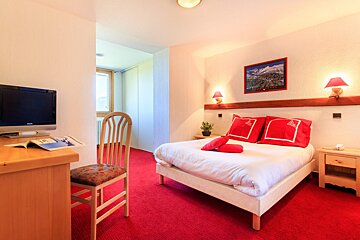 A hotel room with a prominent red carpet, white bed adorned with red pillows, wooden desk, TV, and wall art, lit by warm lamps.
