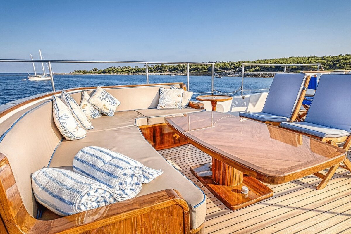 A luxurious yacht deck with cushioned seating, a polished wooden table, blue chairs, overlooking a clear blue sea and distant coastline.