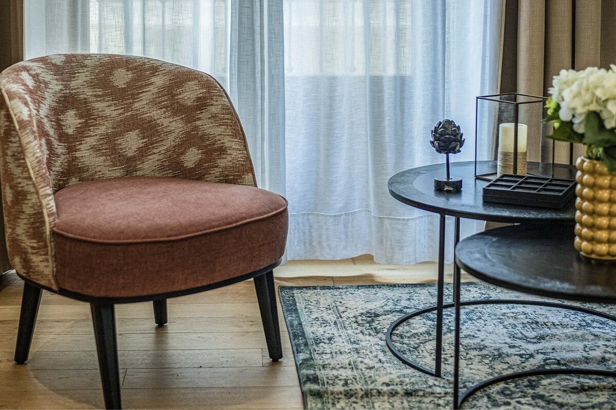 An interior shot featuring a patterned armchair, black nesting tables with decorative items, sheer curtains, and a patterned rug.