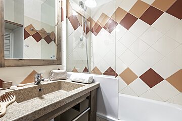 A modern bathroom with a stone sink, wooden-framed mirror, and a tub surrounded by brown and beige diamond-patterned tiles.