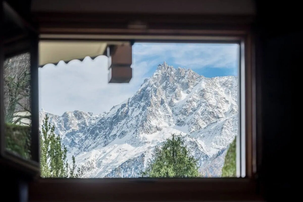 A stunning snow-capped mountain peak is framed by an open window, with glimpses of green trees below under a blue sky.