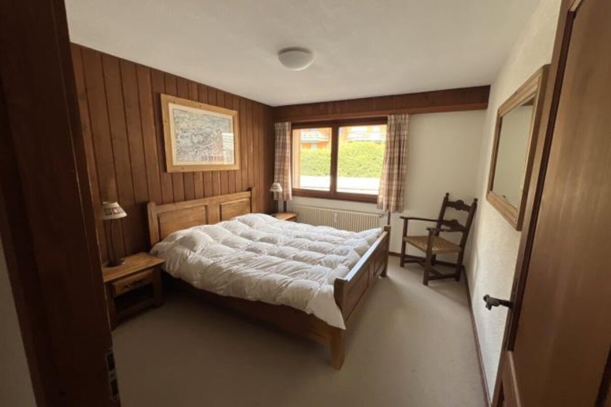 Cozy bedroom featuring wood panel walls, a large wooden bed with a white duvet, a window with outdoor view, and a classic wooden chair.