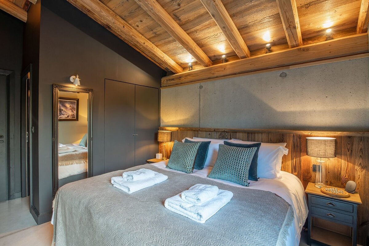 A chic bedroom features a rustic wood-beamed ceiling, dark grey walls, and a concrete accent above a large bed with blue pillows and white towels.