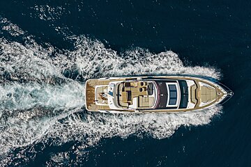 An aerial view of a white luxury yacht with a teak deck speeding across dark blue water, leaving a large foamy white wake.