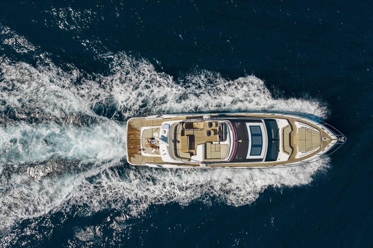 Aerial shot: A white yacht speeds through deep blue water, leaving a large foamy wake. A person relaxes on the stern.