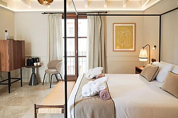 A modern and bright hotel bedroom with a four-poster bed, balcony access, minimalist art, and white bathrobes neatly placed on the bed.