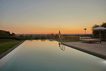 An infinity pool mirrors a serene orange sunset over a marshy landscape. A figure, lounge bed, and umbrella stand by the pool's edge.