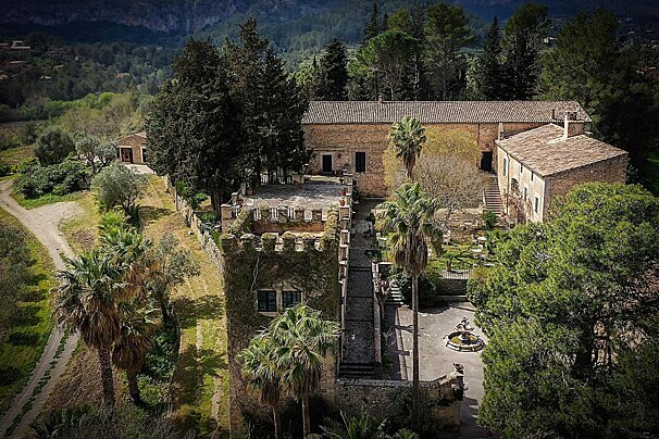Historic stone estate with ivy-clad tower, courtyard fountain, palm trees, and lush surroundings against a mountain backdrop.