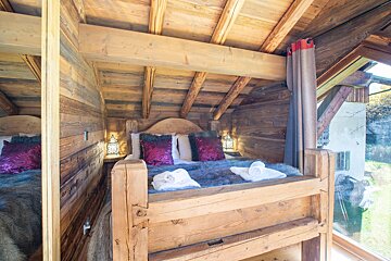 A rustic wooden bedroom features a large bed with purple pillows and a gray furry throw, flanked by ornate lamps. Exposed beams accent the ceiling, and a big window offers an outside view.
