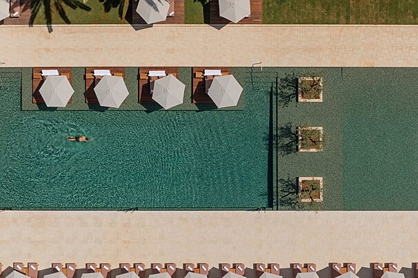 Overhead view of a resort pool with a swimmer, sun loungers and umbrellas, alongside modern landscaping with square tree planters.