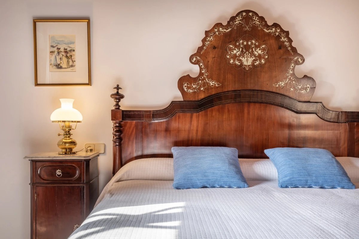 A classic bedroom features an ornate wooden bed, white quilt, blue pillows, a brass lamp on a bedside table, and a framed picture.