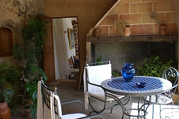 A rustic patio with a blue mosaic table, chairs, and lush plants. Stone walls and a brick fireplace create a cozy setting, with a doorway leading inside.