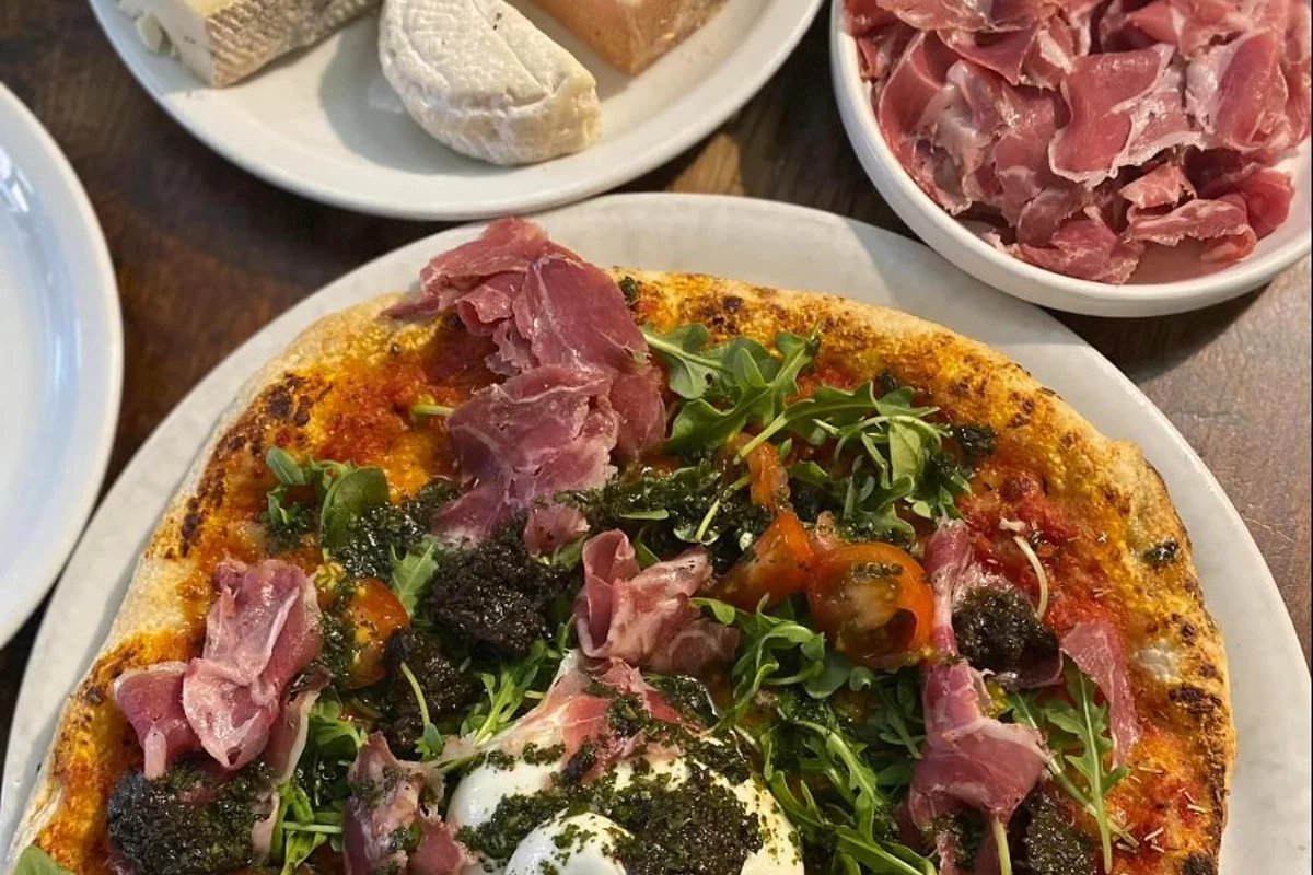 A gourmet pizza topped with prosciutto, arugula, pesto, and burrata, served on a white plate. Artisanal cheeses and more prosciutto are in the background.