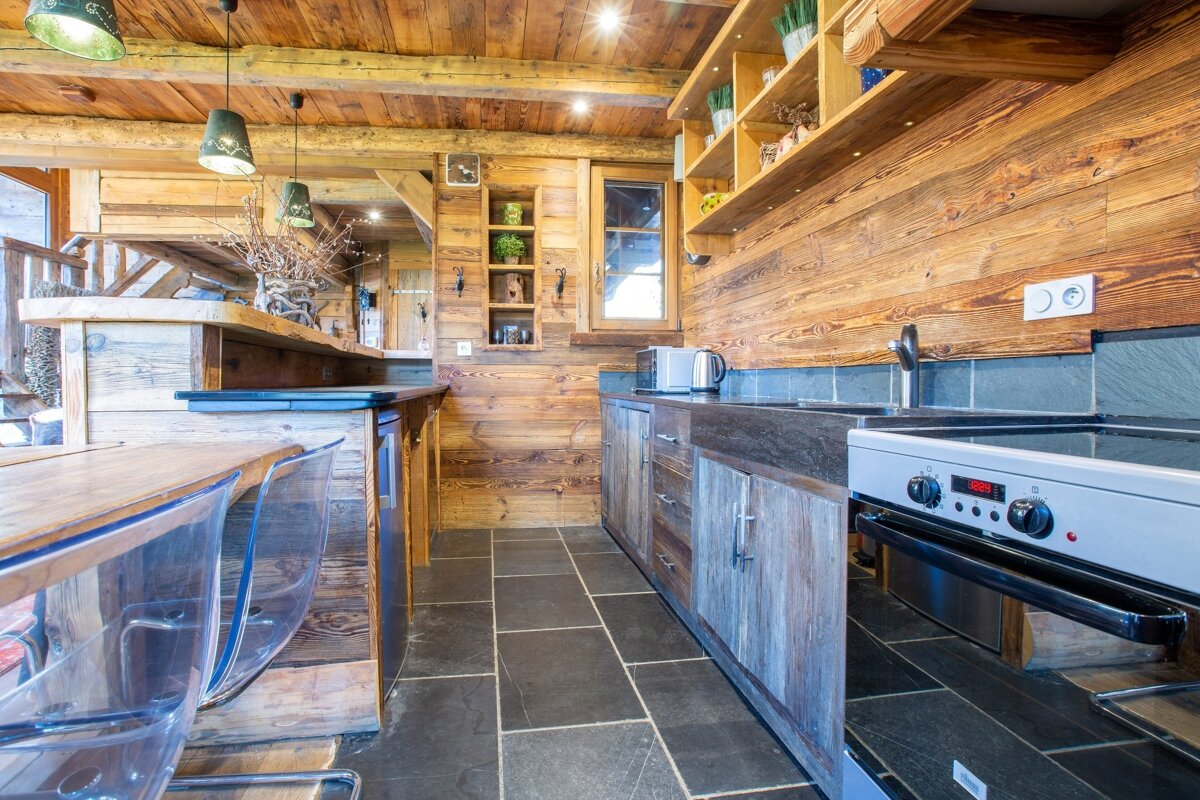 A cozy, chalet-style kitchen featuring extensive wood paneling, dark stone floors/countertops, an oven, microwave, and a bar with clear stools.