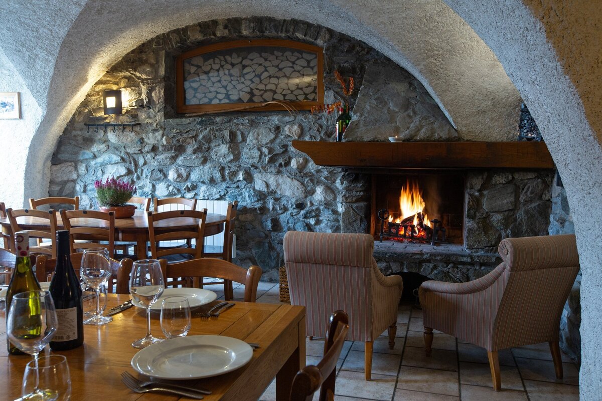 A cozy, rustic dining room with stone walls, arched ceilings, and a crackling fireplace. Tables are set with wine, and armchairs sit by the fire.