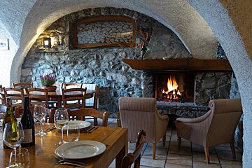 A cozy, rustic dining room with stone walls, arched ceilings, and a crackling fireplace. Tables are set with wine, and armchairs sit by the fire.