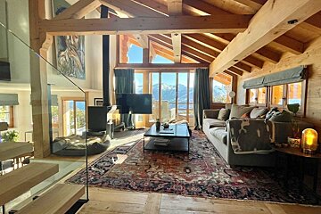 Warm, beamed chalet living room with a fireplace, ornate rug, and panoramic windows revealing snowy mountain vistas.