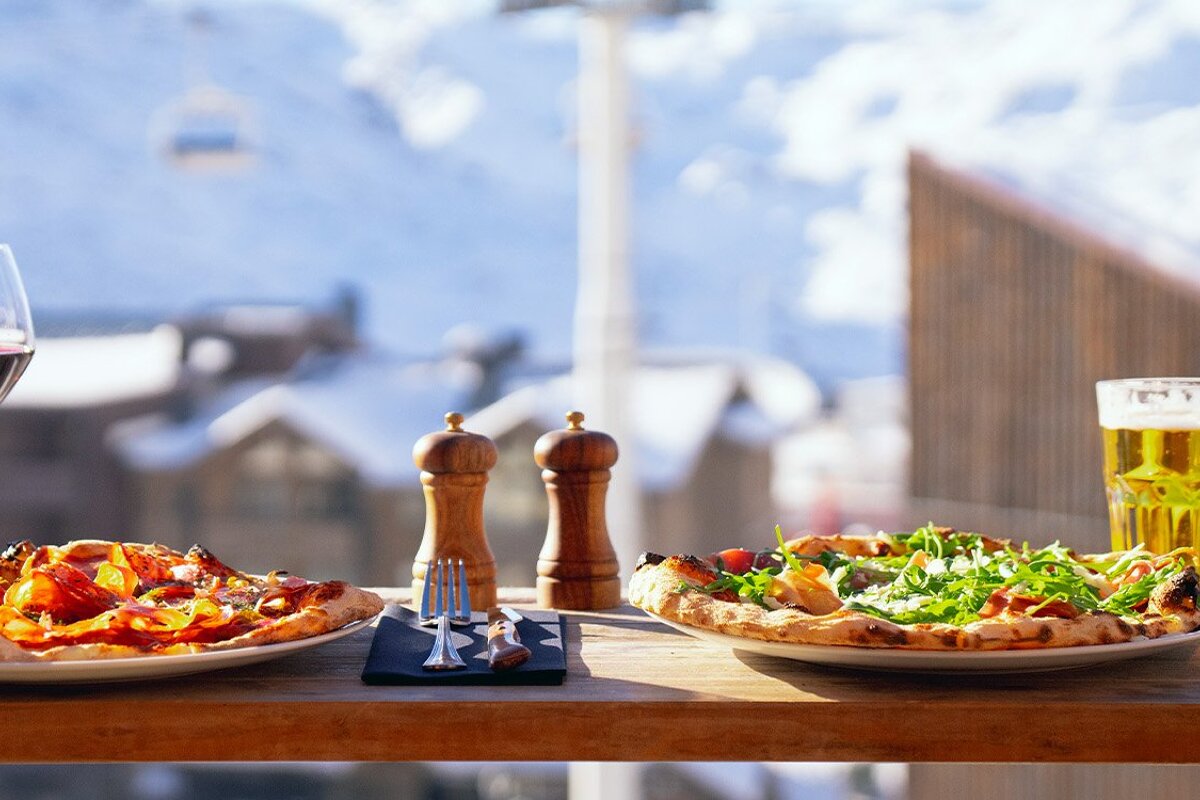 Two pizzas and drinks on a wooden table set against a picturesque snowy mountain backdrop, complete with buildings and a distant ski lift.