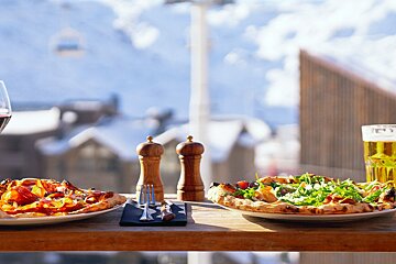 Two pizzas and drinks on a wooden table set against a picturesque snowy mountain backdrop, complete with buildings and a distant ski lift.
