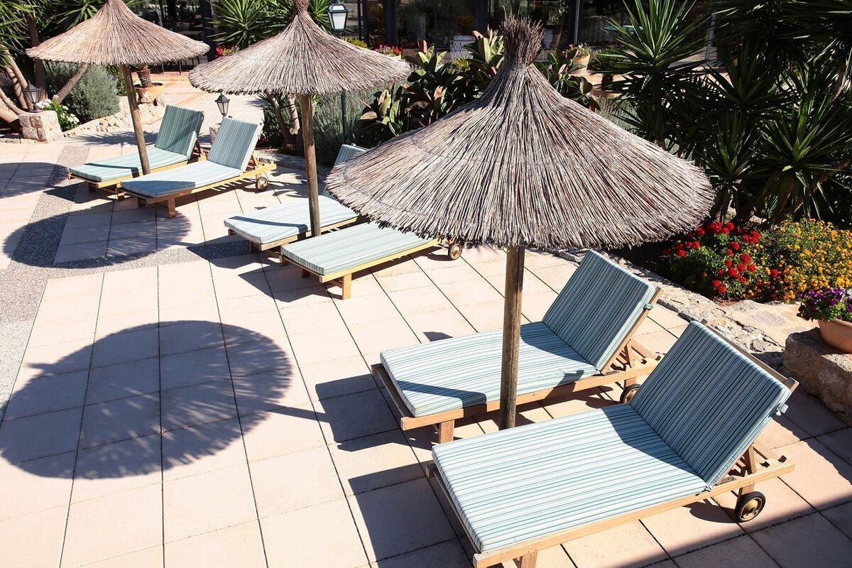 Sunny patio with straw umbrellas casting shadows over lounge chairs with striped cushions, surrounded by lush green plants.