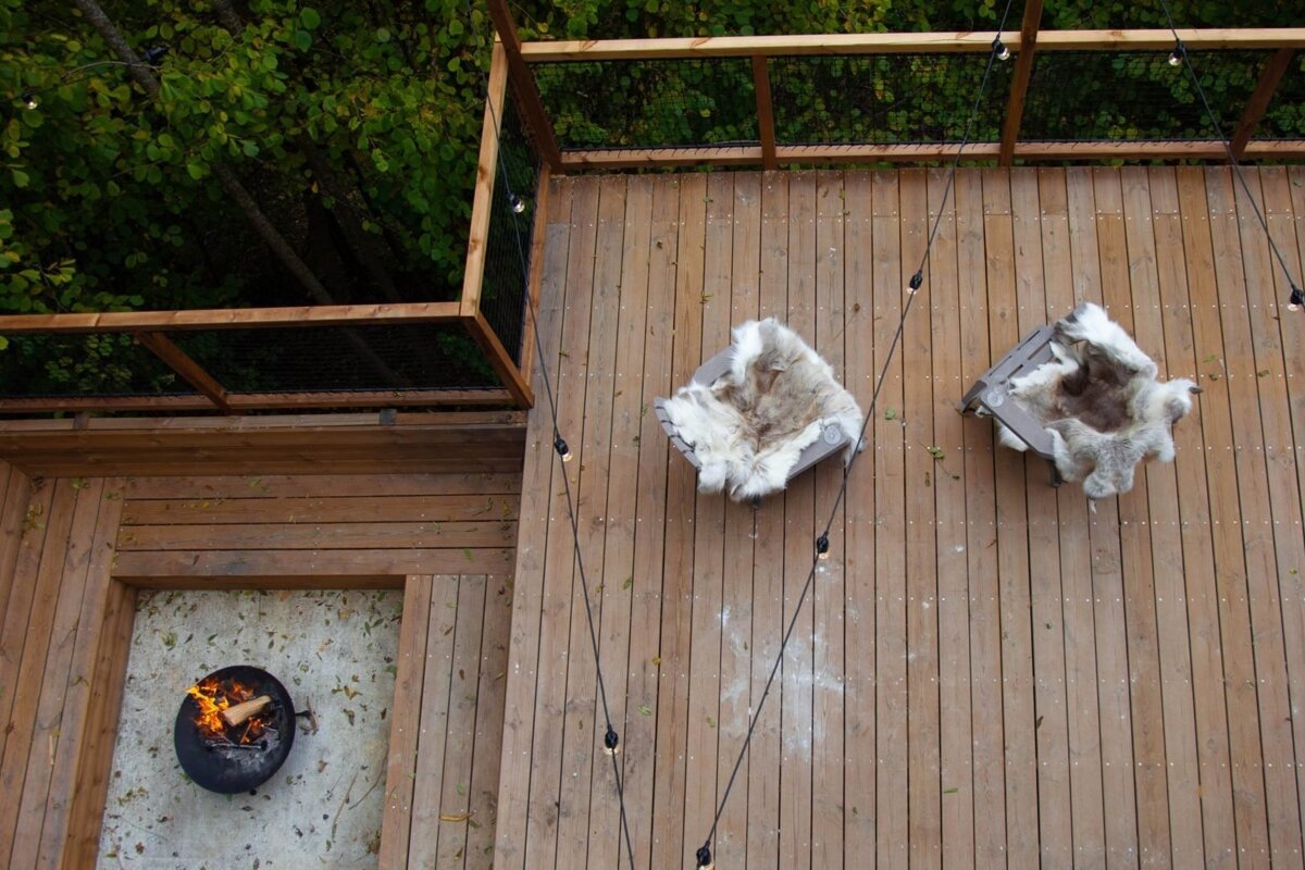 Overhead view of a wooden deck with two furry-covered chairs, a lit fire pit, and string lights. Green trees surround this cozy outdoor setting.