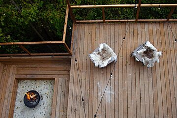 Overhead view of a wooden deck with two furry-covered chairs, a lit fire pit, and string lights. Green trees surround this cozy outdoor setting.
