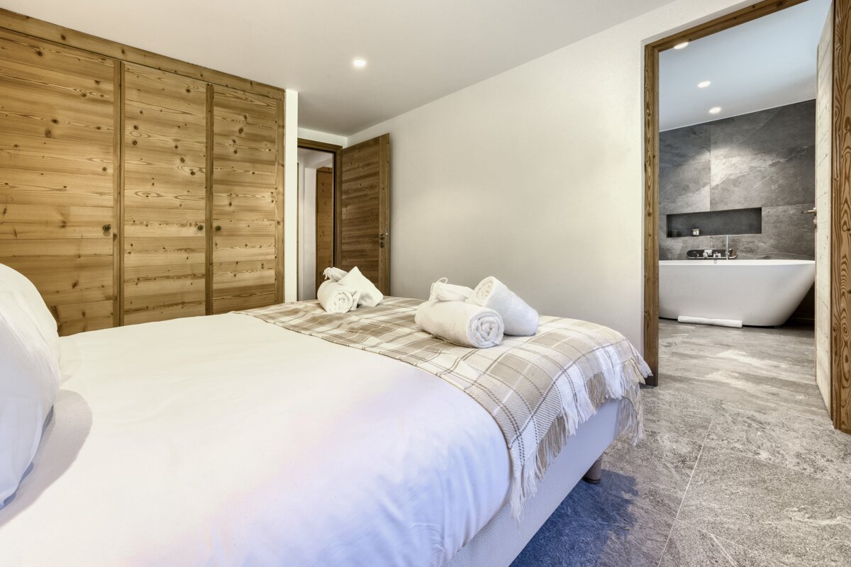 A modern bedroom with a white bed, wood paneling, and a plaid throw. It opens to a sleek bathroom with a freestanding white tub and gray tiles.