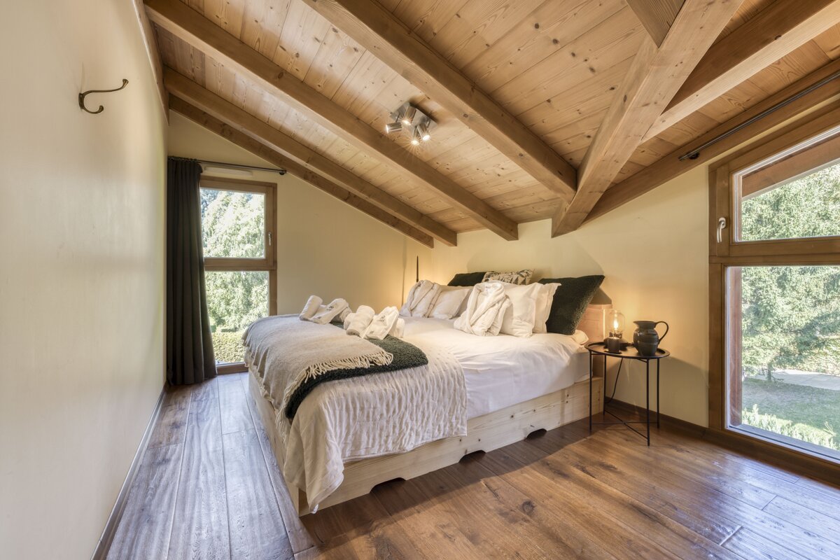 Cozy attic bedroom with a large bed, sloped wooden ceiling and floor, and big windows looking out onto lush greenery.