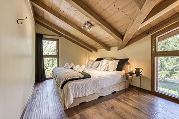 Cozy attic bedroom with a large bed, sloped wooden ceiling and floor, and big windows looking out onto lush greenery.