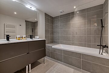 A modern bathroom with gray tiled walls and floor, featuring a double vanity with a large mirror, and a sleek bathtub with an integrated shower.
