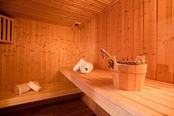 A warm, light-wood sauna interior with wooden benches, rolled white towels, and a traditional wooden bucket with a ladle.