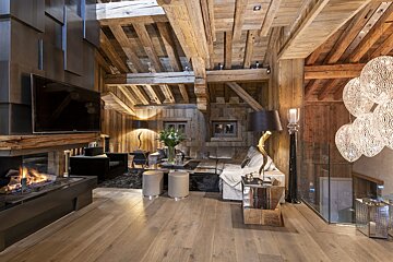 Luxurious chalet living room with exposed wooden beams, a modern fireplace, TV, plush seating, and elegant, ornate lighting fixtures.