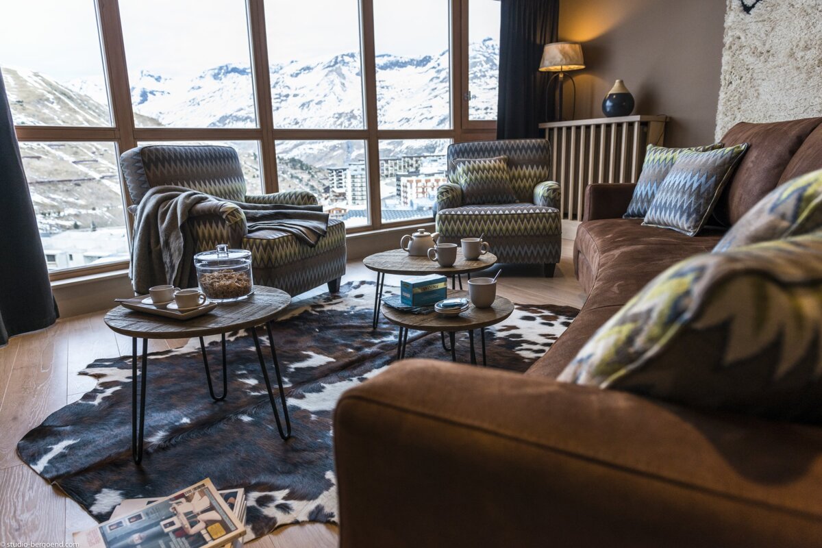 A cozy alpine living room with panoramic snow-capped mountain views, featuring patterned armchairs, a brown sofa, and a cowhide rug.
