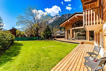 A wooden deck with chairs and tables in front of a house with mountains in the background