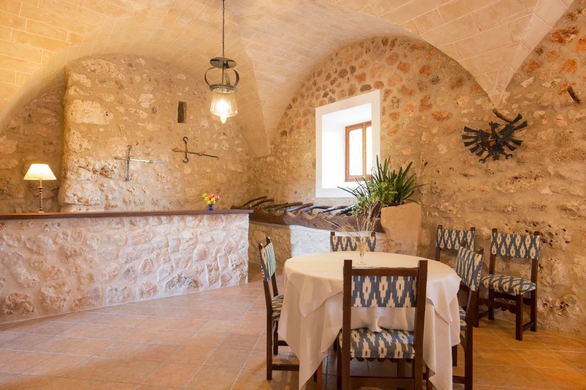 A rustic, vaulted stone room features a dining table with patterned chairs, a bar area, a window, and antique lighting, creating a historic ambiance.