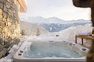 A bubbling hot tub on a snowy patio overlooks majestic, snow-capped mountains. A stone wall is on the left, with wine and a towel nearby.