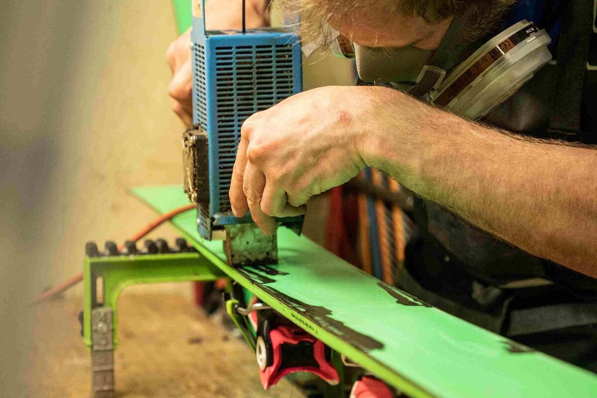 A person wearing a respirator hot waxes a neon green ski, melting wax onto its base with a blue iron for maintenance.