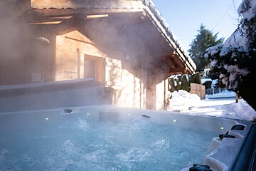 A steaming hot tub bubbles outdoors on a sunny winter day, set against a rustic wooden chalet and snow-covered trees.