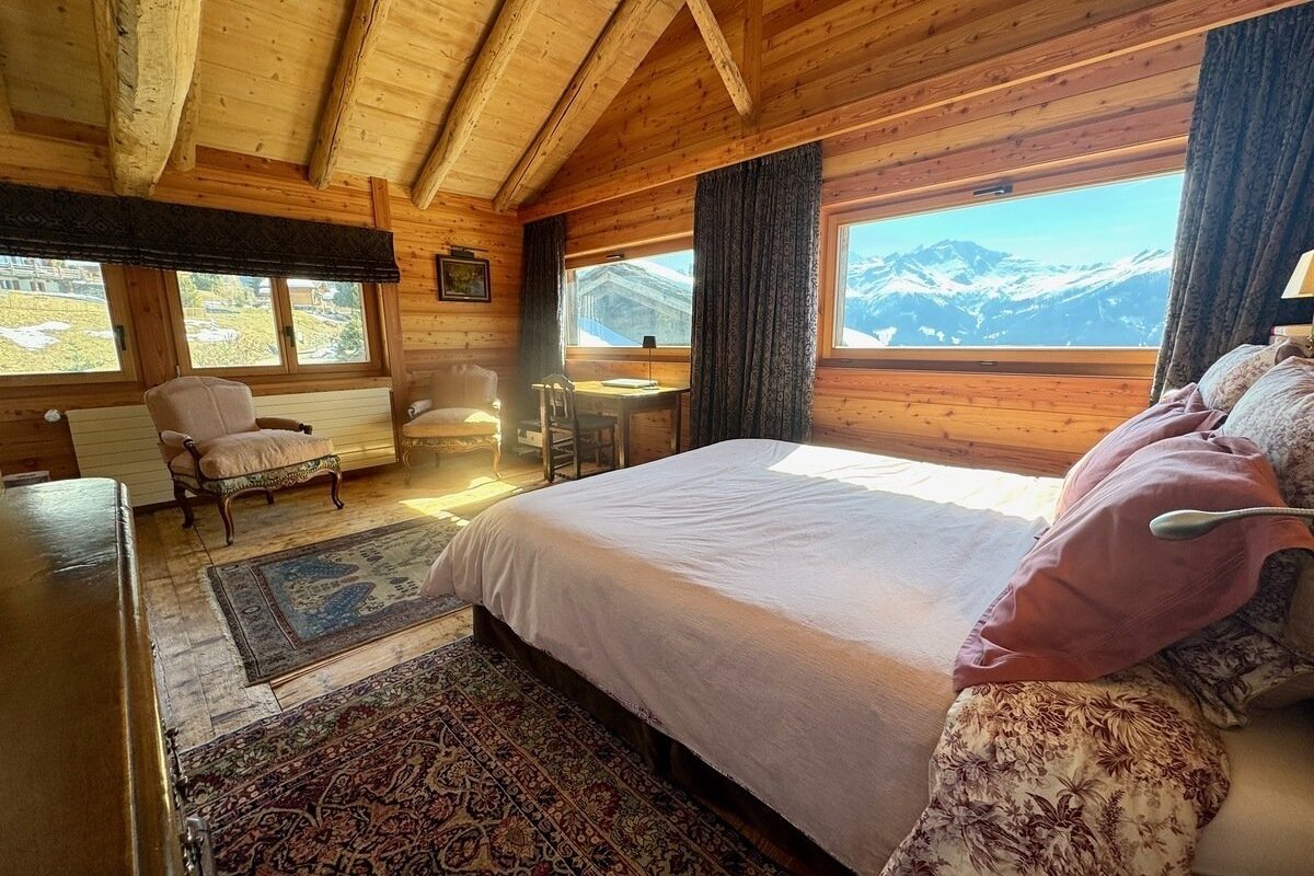 Cozy wooden chalet bedroom with a stunning snowy mountain view from large windows. Features a bed, antique chairs, and a small desk.