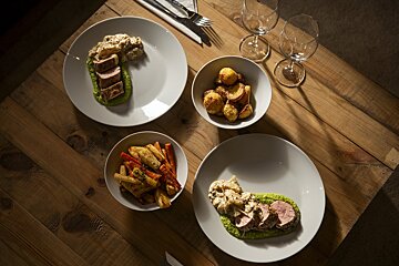 Two plates of sliced meat with green purรฉe, creamy sauce, and roasted root vegetables, alongside roasted potatoes and wine glasses on a rustic wooden table.
