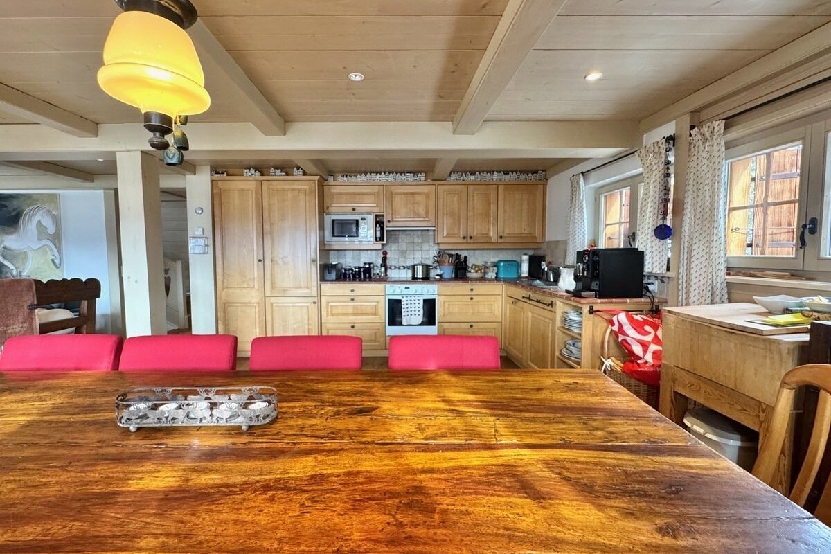 A rustic kitchen and dining area with a large wooden table, red cushioned seats, light wood cabinets, appliances, and beamed ceiling.