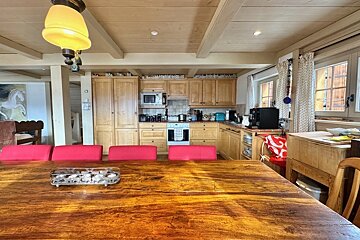 A rustic kitchen and dining area with a large wooden table, red cushioned seats, light wood cabinets, appliances, and beamed ceiling.