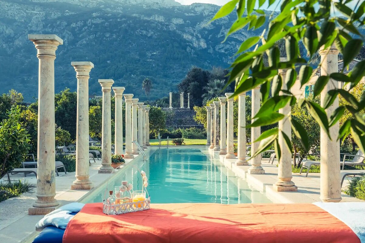 Elegant pool lined with classical columns, mountains in the distance. Red lounge bed & drinks in foreground, surrounded by lush garden foliage.