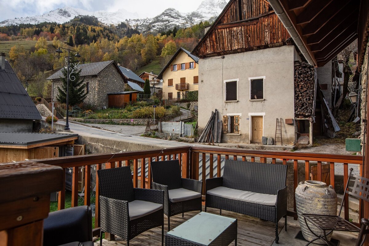 A rustic wooden terrace with patio furniture overlooks a charming mountain village, surrounded by autumn trees and snow-capped peaks under a cloudy sky.