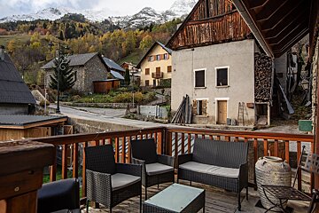 A rustic wooden terrace with patio furniture overlooks a charming mountain village, surrounded by autumn trees and snow-capped peaks under a cloudy sky.
